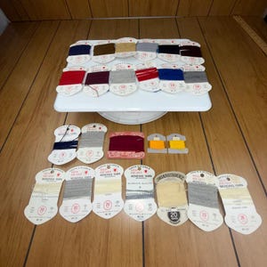 Może przedstawiać: Kolekcja szpulek z przędzy do cerowania w stylu vintage w różnych kolorach, w tym czerwonym, niebieskim, szarym i brązowym. Szpulki ułożone są na białym obrotowym talerzu i drewnianej powierzchni. Etykiety z napisem "Red Heart Mending Yarn".