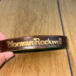May include: A brown leather bracelet with the name "Norman Rockwell" embossed in gold lettering. The bracelet is curved and appears to be new.