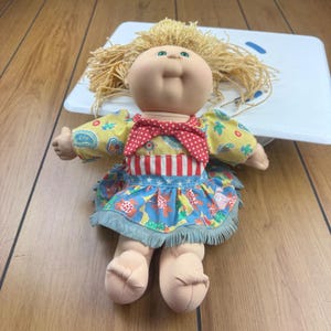 Könnte beinhalten: Vintage Cabbage Patch Kid Puppe mit blonden Garnhaaren, rundem Gesicht und grünen Augen. Die Puppe trägt ein gelb-blaues Blumenkleid mit roter Schleife und eine blau-weiß gestreifte Schürze. Ein klassisches Sammlerspielzeug.