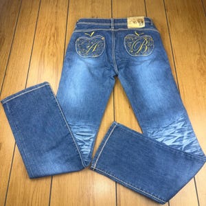 May include: Blue denim jeans with gold embroidered apple designs on the back pockets. The jeans have a classic five-pocket design, a button and zipper closure, and white stitching. The jeans are laid flat on a wooden surface.