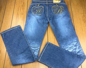 May include: Blue denim jeans with gold embroidered apple designs on the back pockets. The jeans have a classic five-pocket design, a button and zipper closure, and white stitching. The jeans are laid flat on a wooden surface.