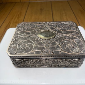 May include: A silver-coloured, rectangular jewellery box with detailed floral and vine patterns. The box has a central oval accent and ornate edges. The design is embossed, creating a textured surface. The box is likely made of metal.
