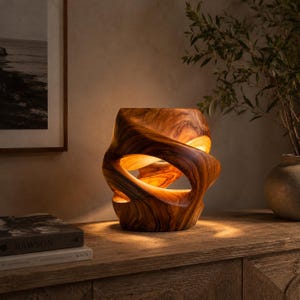 Könnte beinhalten: Eine Tischlampe aus Holz mit einem einzigartigen, organischen Design. Die Lampe ist aus Holz geschnitzt und weist fließende, geschwungene Linien und offene Räume auf, durch die das Licht scheinen kann. Der warme, bernsteinfarbene Schein erzeugt ein gemütliches Ambiente. Die Lampe ist etwa 30 cm hoch.