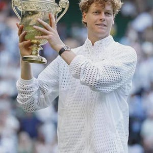 May include: A person with curly hair wearing a white long-sleeved jacket with a textured mesh pattern on the upper chest and sleeves, holding a large, ornate gold-colored trophy with two handles above their head while wearing a wristwatch.