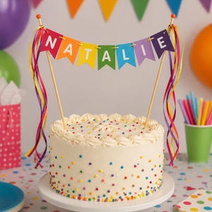 May include: A round, white frosted cake decorated with colourful sprinkles and a banner spelling "NATALIE". The banner is in rainbow colours and is held up by wooden sticks. The cake is on a white stand.