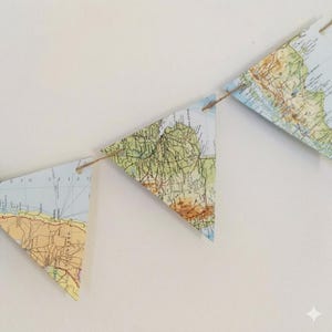Peut inclure: Une guirlande décorative composée de fanions triangulaires en papier arborant des détails de cartes géographiques imprimées dans des tons de vert, de beige et de bleu. Les fanions sont reliés par une fine cordelette en fibre naturelle.