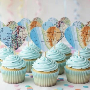 May include: A dozen cupcakes with light blue frosting and heart-shaped map toppers. The toppers are attached to wooden sticks. The cupcakes are in light blue paper cups and are decorated with colorful confetti.