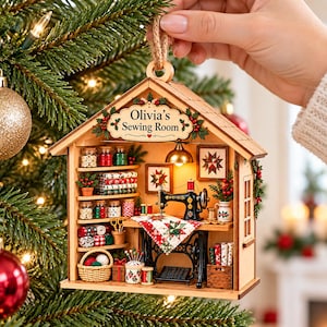 Pode incluir: Um enfeite de Natal de madeira em forma de casa com um interior de sala de costura em miniatura, incluindo uma máquina de costura preta, prateleiras com tecidos, potes de botões, uma cesta de lã e o letreiro Olivia's Sewing Room.