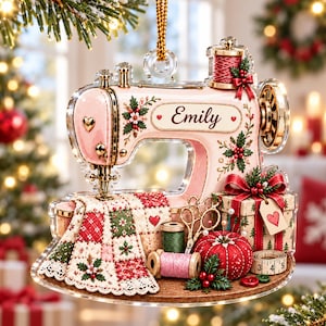 May include: A clear acrylic ornament shaped like a vintage sewing machine in light pink with gold accents. It features the name Emily, a red and green quilted fabric, a spool of thread, gold scissors, a red pincushion, and a gift box with a heart tag.
