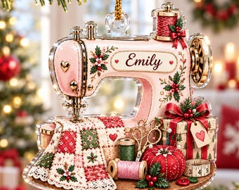 May include: A clear acrylic ornament shaped like a vintage sewing machine in light pink with gold accents. It features the name Emily, a red and green quilted fabric, a spool of thread, gold scissors, a red pincushion, and a gift box with a heart tag.