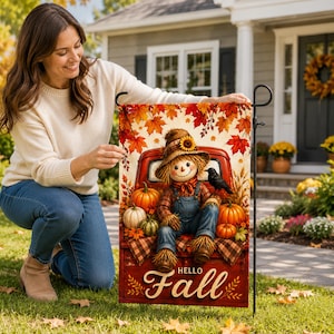 May include: A rectangular garden flag featuring a scarecrow sitting in the bed of a red truck surrounded by pumpkins and autumn leaves. The text "HELLO Fall" is printed in white script at the bottom. A black crow sits on the scarecrow's shoulder.