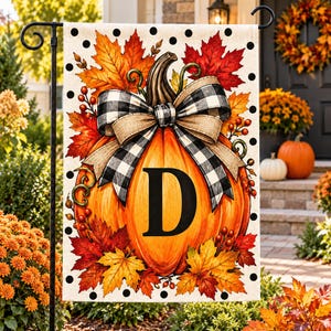 May include: A rectangular garden flag featuring a large orange pumpkin with a black letter D in the center. The pumpkin is adorned with a black and white buffalo plaid bow, surrounded by red and orange autumn leaves, berries, and black polka dots.