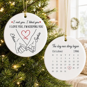 May include: Two round white ceramic ornaments with gold hanging strings. One features a line drawing of hands holding pinky fingers, a red heart, and the text I met you, I liked you, I LOVE YOU, I'M KEEPING YOU with names Ethan and Sophia. The other shows a calendar for October 1986 with the 25th circled in red and the text The day our story began.