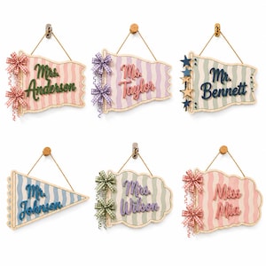 May include: Six hanging wooden signs with wavy striped backgrounds in pastel colors. Each sign features a personalized name in raised lettering, decorated with fabric bows or wooden stars, and suspended by a thin cord from a small wall hook.