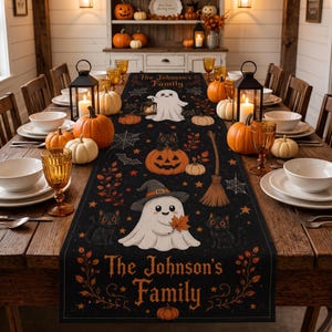 May include: A black rectangular table runner featuring a Halloween theme with ghosts, black cats, pumpkins, spiderwebs, and a broomstick. The text "The Johnson's Family" is printed at both ends of the runner, which is set on a wooden dining table.
