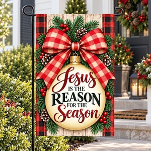 May include: A rectangular garden flag featuring a cream-colored Christmas ornament with the text Jesus is the reason for the season. The ornament is topped with a large red and white buffalo plaid bow, green pine branches, and red berries.