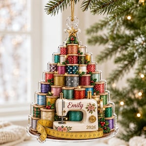 May include: A Christmas tree-shaped ornament made of stacked colorful thread spools, topped with a gold star. At the base sits a white sewing machine with the name Emily and the year 2026 printed on it, wrapped in a yellow measuring tape.