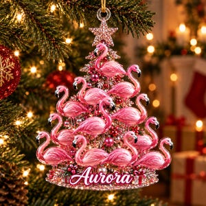 May include: A Christmas tree-shaped ornament featuring multiple pink flamingo figures arranged in tiers. The ornament is topped with a glittery star and includes the name Aurora in white script lettering at the base, accented with small red berries.