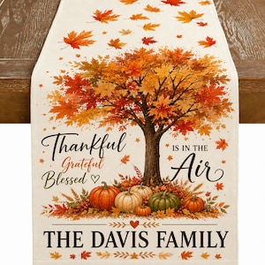 May include: A cream-colored table runner featuring a large autumn tree with orange and red leaves, surrounded by pumpkins and wheat. The text reads: Thankful Grateful Blessed is in the air, and at the bottom, it says: THE DAVIS FAMILY.