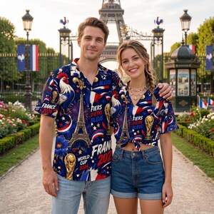 May include: A couple wearing matching blue button-down shirts with a French theme. The shirts feature the Eiffel Tower, a rooster, and the text "France World Cup 2026" in red, white, and blue. They are standing in front of the Eiffel Tower.