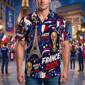 May include: A short-sleeved button-down shirt with a navy blue background, featuring a design of the Eiffel Tower, a rooster, and the words "France 2026." The shirt also includes the words "Fiers d'&ecirc;tre Bleus" and images of the World Cup trophy.