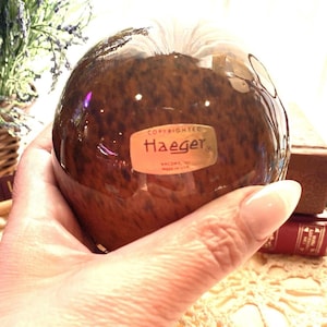 May include: A brown, glossy decorative apple with a speckled pattern. The apple has a gold label that reads "Haeger" and "Copyrighted". The apple is held in a hand.
