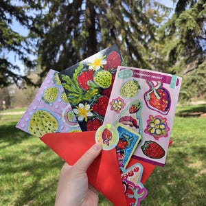 Peut inclure: Une enveloppe en papier rouge contient plusieurs cartes et autocollants avec des illustrations de fraises. Les cartes présentent des peintures détaillées et des planches d'autocollants avec divers motifs de fraises et le texte "Strawberry Delight". Un autocollant holographique de fraise est également visible.