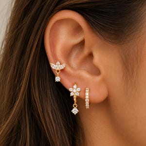 May include: A set of three gold-tone earrings featuring clear cubic zirconia stones. The top earring is a lotus-shaped stud with a dangling stone, the middle is a flower-shaped hoop with a dangling stone, and the bottom is a small pavé hoop.