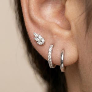 May include: Close-up of three earrings on an ear. One is a leaf-shaped stud with multiple clear stones. The second is a small hoop with clear stones, and the third is a plain silver hoop. Jewelry.