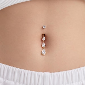 May include: A gold-toned navel piercing featuring a small round clear gemstone at the top and a dangling vertical chain of three graduated pear-shaped clear gemstones. The jewelry is set in a metallic gold-colored base and rests in a navel.