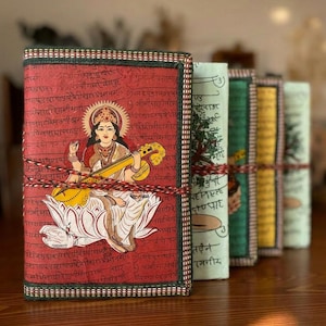 Può includere: Una fila di diari decorativi e colorati. Il primo diario presenta una copertina rossa con un'illustrazione di una divinità che suona uno strumento a corde. Gli altri diari hanno copertine verdi, gialle e verde chiaro. Ogni diario è rilegato con un cordoncino decorativo.