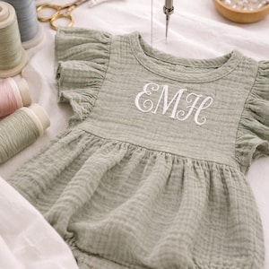 Könnte beinhalten: Hellgrüner Baby-Strampler mit Rüschenärmeln und gerafftem Rock. Der Strampler hat die gestickten Initialen "EMH" in weißer Schrift. Nähmaschine, Garnrollen und Schere im Hintergrund, was auf Handarbeit hindeutet.