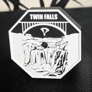 May include: A black and white octagon-shaped object with the words "TWIN FALLS" at the top. The design features a bridge, waterfalls, and a stylized landscape. A small black knob is in the center.