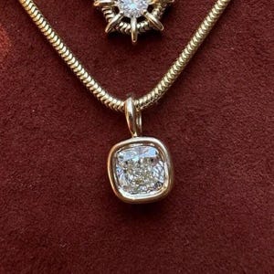 May include: A gold chain necklace featuring a square-shaped pendant with a central clear faceted stone. A second, partially visible pendant with a starburst design and a central clear stone is positioned above the main square pendant.