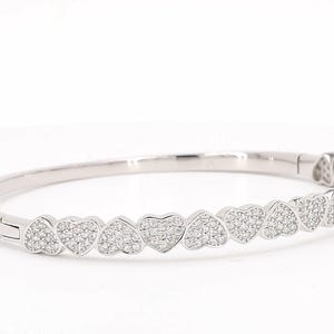 May include: A silver-toned hinged bangle bracelet featuring a continuous row of heart-shaped links. Each heart link is encrusted with small, clear, sparkling stones across its surface. The bracelet has a secure clasp mechanism on the side.