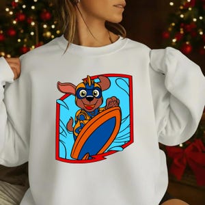 Op de afbeelding: Witte sweatshirt met een cartoonhond die surft. De hond draagt een blauw en oranje superheldenoutfit en houdt een surfboard vast. Het ontwerp staat in een rood omrand schild tegen een blauwe achtergrond.