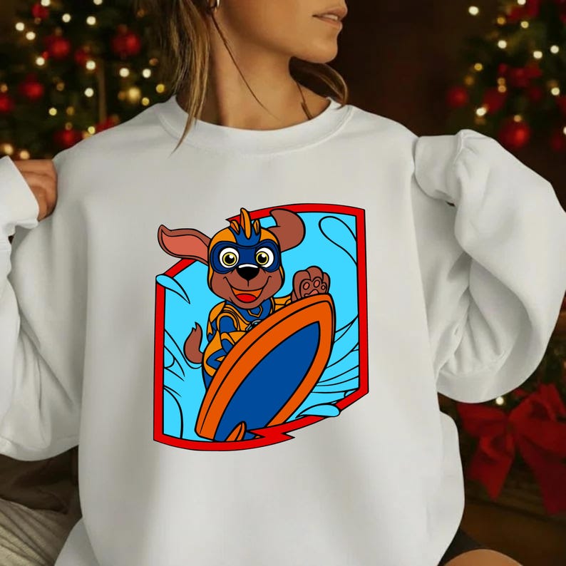Op de afbeelding: Witte sweatshirt met een cartoonhond die surft. De hond draagt een blauw en oranje superheldenoutfit en houdt een surfboard vast. Het ontwerp staat in een rood omrand schild tegen een blauwe achtergrond.