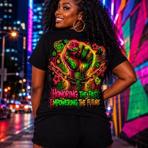 Könnte beinhalten: Ein schwarzes T-Shirt mit einer leuchtenden Neon-Grafik einer erhobenen Faust, die Ketten durchbricht und mit komplexen Mustern verziert ist. Der Text "HONORING THE PAST EMPOWERING THE FUTURE" steht in fetten, bunten Buchstaben auf dunklem Grund.