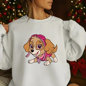 Op de afbeelding: Witte sweatshirt met een cartoonafbeelding van Skye van Paw Patrol. Het personage is afgebeeld in roze en bruine kleuren, met een roze pilotenhelm en vest. Het ontwerp is gecentreerd op de voorkant van de sweatshirt.