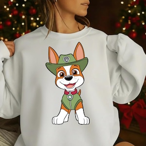 Op de afbeelding: Witte sweatshirt met een cartoon hond karakter met een groene hoed en vest. De hond heeft bruin en wit haar, een glimlachende uitdrukking en een badge op zijn hoed en vest. De achtergrond is wazig met kerstversieringen.