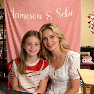 May include: Two women smile in front of a pink backdrop. The backdrop has the text "Jameson & Sebe" in white script. One woman wears a white dress, and the other wears a red and white striped shirt.