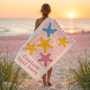 Peut inclure: Une serviette de plage avec un motif à carreaux roses et blancs, ornée d'étoiles de mer jaunes, bleues et roses. Le texte "Custom text here" est en rose. Le fond montre une scène de plage avec un coucher de soleil.