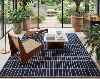 Puede incluir: Un salón moderno con una alfombra azul marino con un patrón rectangular beige. Dos sillas de madera con asientos tejidos frente a una mesa de centro de mármol. La habitación está llena de plantas y tiene vistas a un jardín.