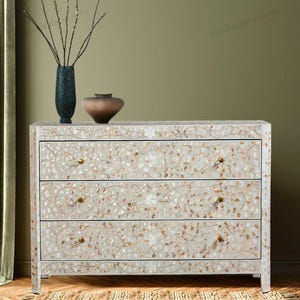 May include: A rectangular chest of drawers featuring three drawers with an intricate floral inlay pattern made of iridescent shell pieces. The chest has a light cream finish with gold-toned hardware knobs on each drawer front.