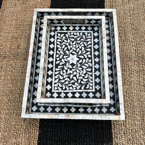 May include: A set of two rectangular nesting trays featuring a black base with intricate white mother-of-pearl inlay. The design includes a central floral motif surrounded by a geometric diamond border and a solid mother-of-pearl frame edge.