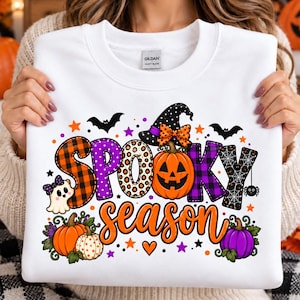 Puede incluir: Una sudadera blanca de cuello redondo con un gráfico colorido que incluye el texto "SPOOKY season". El diseño presenta una calabaza de Halloween, un fantasma, murciélagos, una araña, calabazas y estrellas con patrones en naranja, morado, negro y blanco.