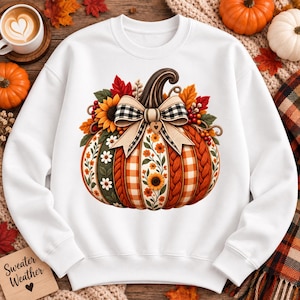 May include: A white long-sleeve sweatshirt featuring a large central graphic of a pumpkin decorated with sunflowers, daisies, and autumn leaves. The pumpkin has alternating panels of orange plaid, cable knit, and floral patterns with a plaid bow.