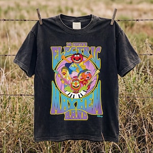 Puede incluir: Una camiseta negra de manga corta con un gráfico colorido de la banda Dr. Teeth and the Electric Mayhem. El diseño incluye a los miembros de la banda, un fondo circular y el texto Dr. Teeth's Electric Mayhem Band en morado y amarillo.