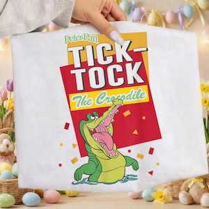 Op de afbeelding: Een wit t-shirt met een grafische print met de tekst Peter Pan TICK-TOCK The Crocodile. Het ontwerp bevat een grote groene krokodil met een open bek, geplaatst tegen een rode en gele rechthoekige achtergrond met confetti-vormen.