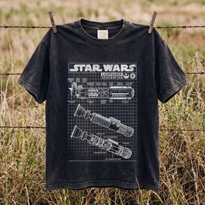 Puede incluir: Una camiseta negra de manga corta con un estampado gráfico blanco de esquemas de sables de luz de Star Wars. El diseño incluye diagramas técnicos de componentes de sables de luz sobre un fondo de cuadrícula con el texto Star Wars Lightsaber Schematics.